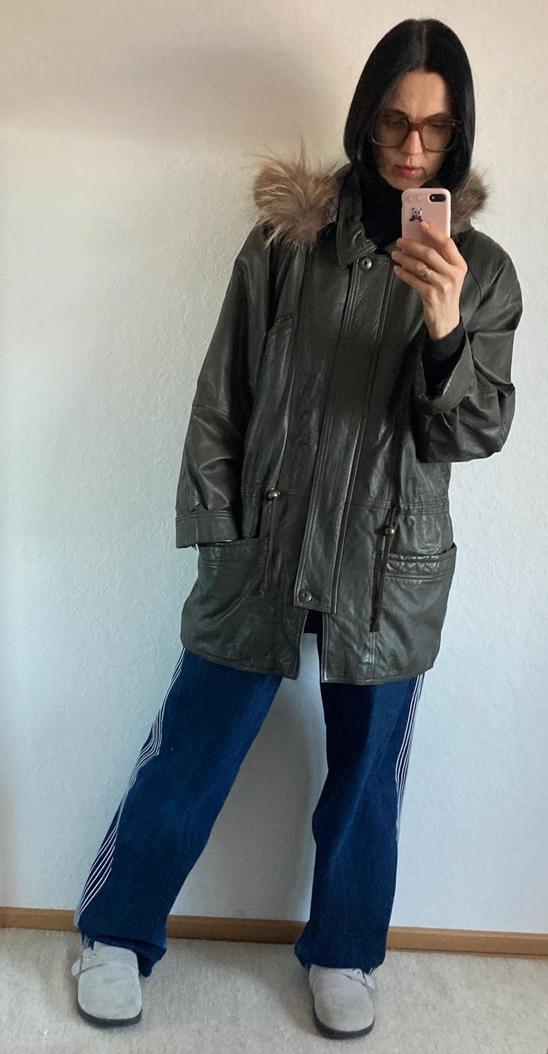 May include: Dark green leather jacket with a fur-trimmed hood. The jacket has a zip and button closure, two front pockets, and a drawstring waist. The person is wearing blue jeans with white stripes and gray shoes.