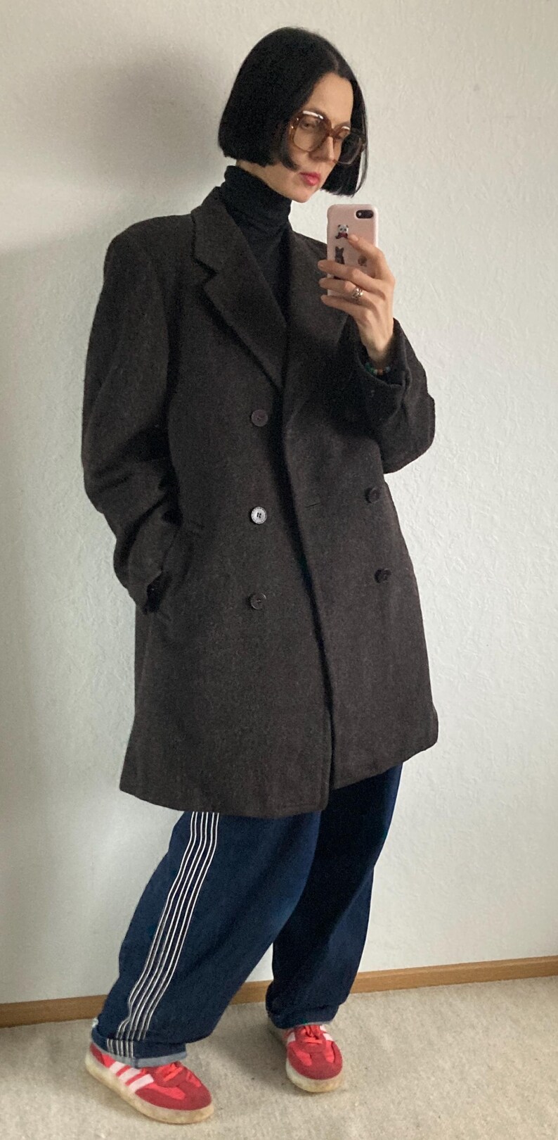 May include: A dark brown double-breasted coat with large buttons. The coat is worn with a black turtleneck, blue sweatpants with white stripes, and red sneakers with white stripes.