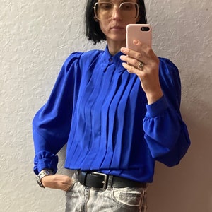 Può includere: Una donna indossa una blusa blu reale a maniche lunghe e plissettata con colletto. Indossa una cintura nera e jeans grigi.