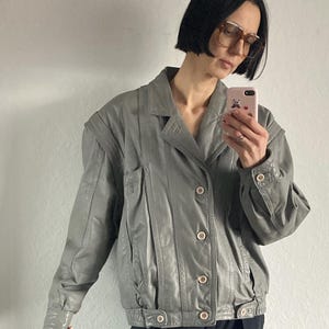 May include: A gray leather jacket with a classic design. The jacket features a collared neckline, button closures, and a pleated front. The jacket has a slightly oversized fit and is a versatile outerwear piece.