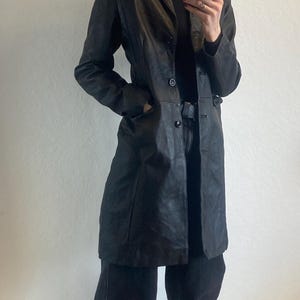 Puede incluir: Un abrigo largo de cuero negro con cierre de botones, usado sobre un cuello alto negro y pantalones vaqueros oscuros. El abrigo tiene un diseño clásico y entallado. El atuendo se completa con zapatos negros.