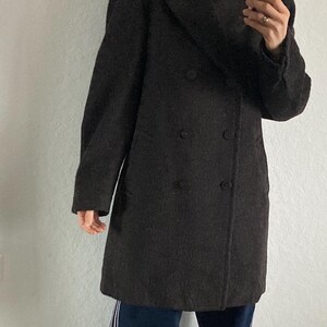 May include: A dark brown double-breasted coat. The coat is worn over a black turtleneck and paired with dark blue pants with white stripes down the side. Red sneakers complete the look.