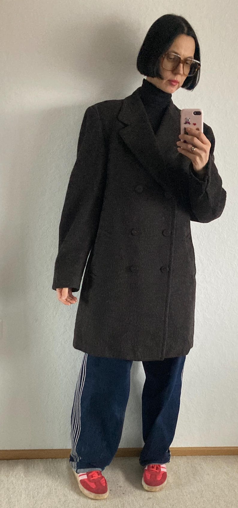 May include: A dark brown double-breasted coat. The coat is worn over a black turtleneck and paired with dark blue pants with white stripes down the side. Red sneakers complete the look.
