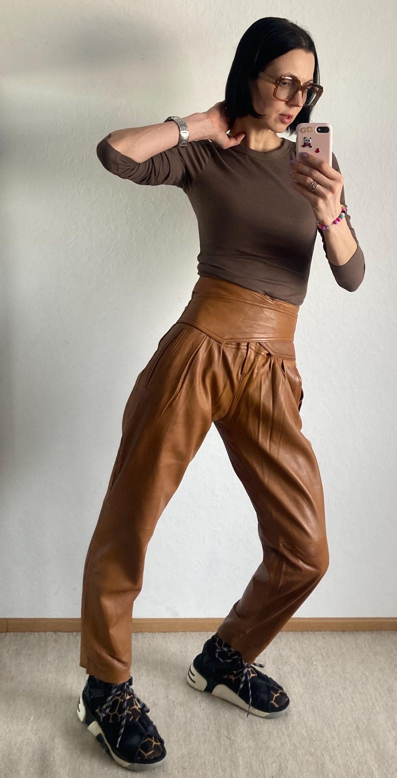 May include: A person wearing a brown long-sleeve shirt and high-waisted, brown leather pants. The person is also wearing black and white sneakers with a leopard print design.