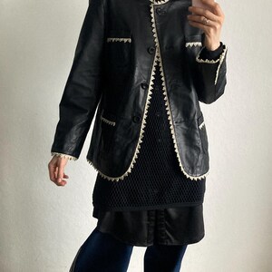 May include: Black leather jacket with white trim, worn over a black top and skirt. The jacket has a classic design with button closures and pocket details. The outfit is paired with dark blue pants and yellow sneakers.