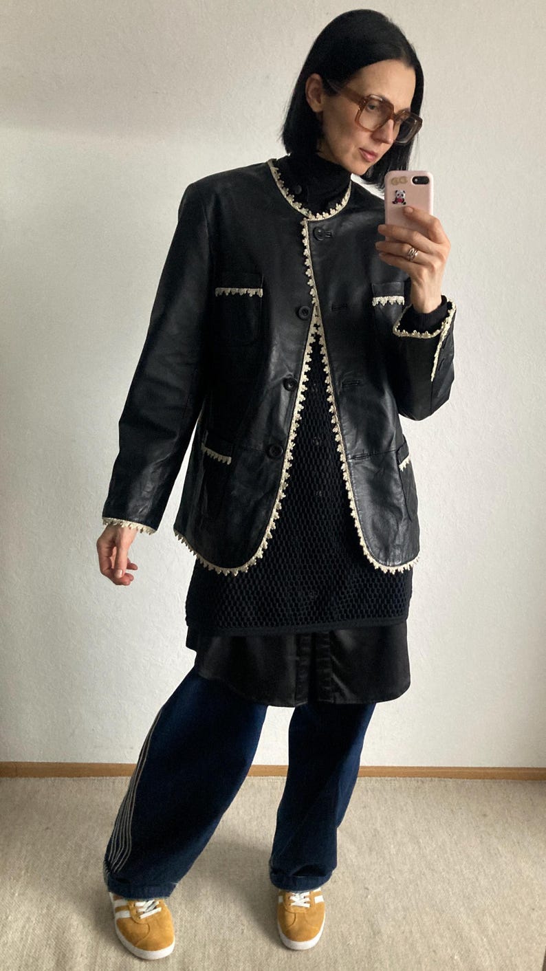 May include: Black leather jacket with white trim, worn over a black top and skirt. The jacket has a classic design with button closures and pocket details. The outfit is paired with dark blue pants and yellow sneakers.