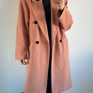 May include: A double-breasted, peach-colored coat with black buttons. The coat is worn over a black turtleneck and paired with dark blue pants and red sneakers. The outfit is set against a plain white wall.
