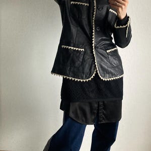 May include: Black leather jacket with cream-colored trim and a matching collar. The jacket is paired with a black top, dark blue pants, and tan sneakers with white stripes. The outfit is a mix of textures and styles.