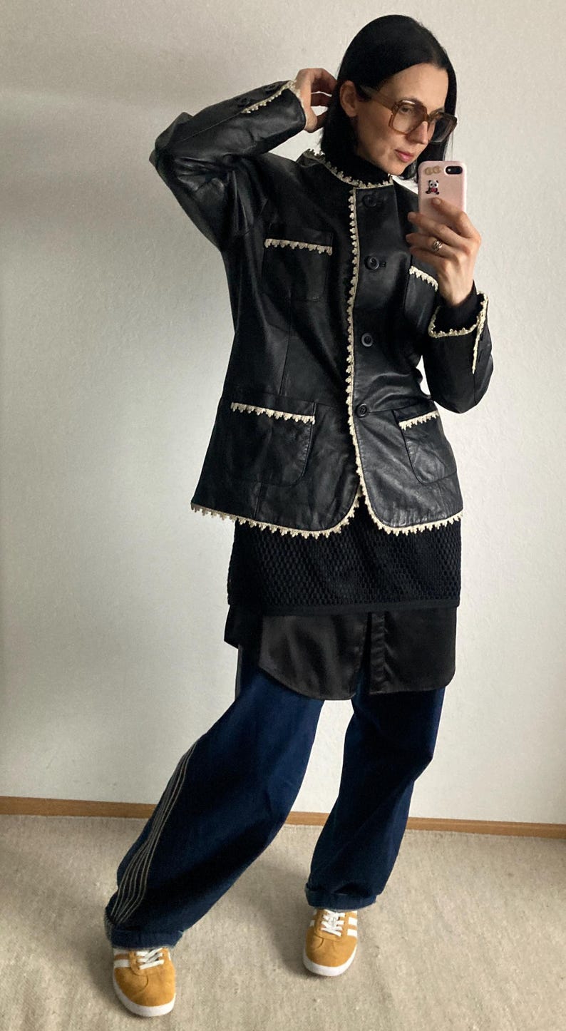 May include: Black leather jacket with cream-colored trim and a matching collar. The jacket is paired with a black top, dark blue pants, and tan sneakers with white stripes. The outfit is a mix of textures and styles.