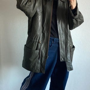 May include: An olive green leather jacket with a fur-lined hood. The jacket has a zipper closure, pockets, and a drawstring waist. The person is wearing dark blue jeans and gray shoes.