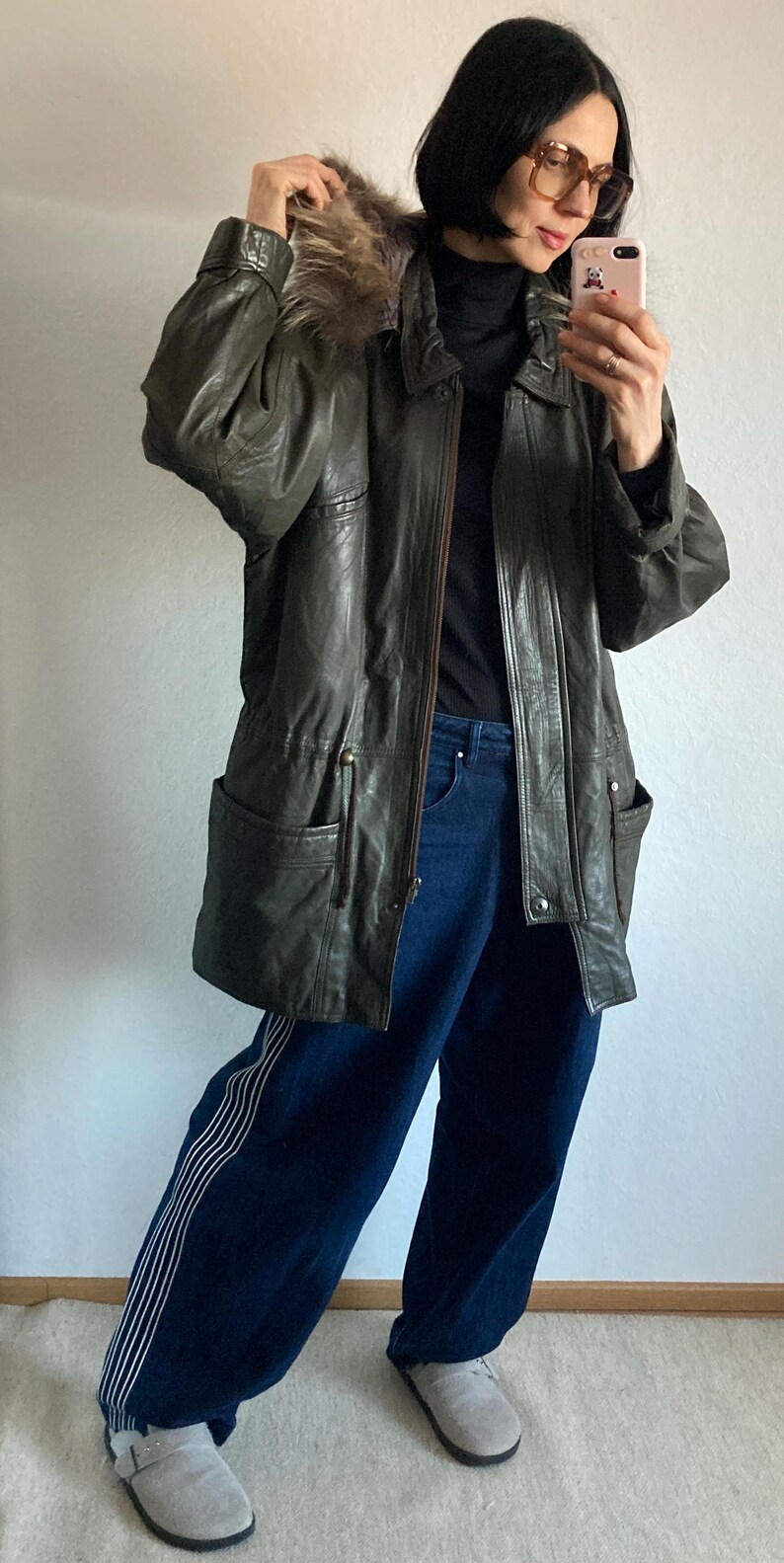 May include: An olive green leather jacket with a fur-lined hood. The jacket has a zipper closure, pockets, and a drawstring waist. The person is wearing dark blue jeans and gray shoes.