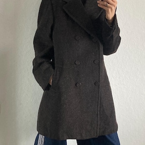 May include: A dark brown double-breasted coat with two rows of buttons. The coat is worn over a black turtleneck. The person is wearing dark blue jeans with white stripes and red sneakers.