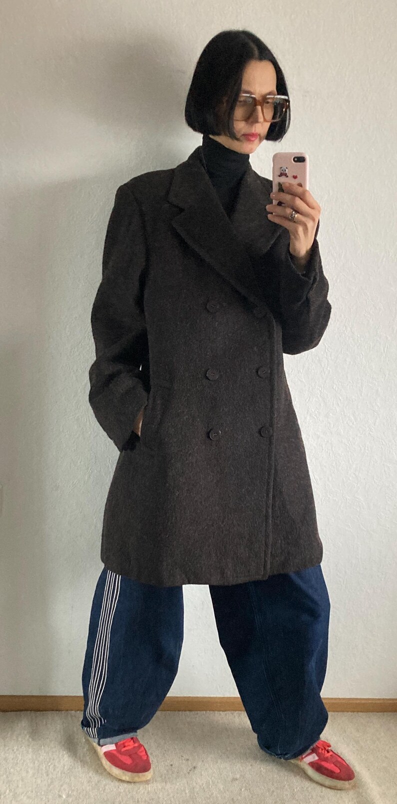 May include: A dark brown double-breasted coat with two rows of buttons. The coat is worn over a black turtleneck. The person is wearing dark blue jeans with white stripes and red sneakers.