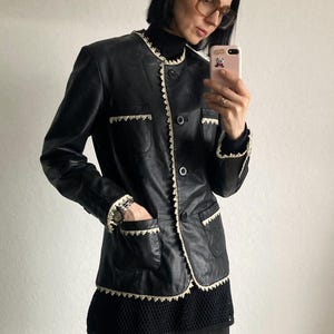 May include: Black leather jacket with cream-colored trim and pockets. The jacket has a round collar and button closure. The trim is a decorative, scalloped design. The jacket is worn over a black top and blue jeans.