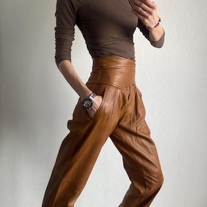 May include: A brown long-sleeve top and high-waisted leather pants in a warm brown hue. The pants have a unique pleated design. The outfit is paired with patterned sneakers. A silver watch is visible on the wrist.