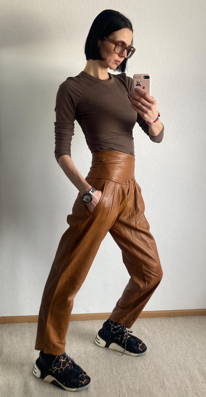 May include: A brown long-sleeve top and high-waisted leather pants in a warm brown hue. The pants have a unique pleated design. The outfit is paired with patterned sneakers. A silver watch is visible on the wrist.