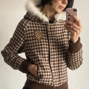 Puede incluir: Chaqueta marrón y crema con estampado de pata de gallo y capucha forrada de piel sintética. La chaqueta presenta cuello y puños de punto marrón, botones dorados y un emblema dorado en el pecho. La chaqueta se lleva sobre una falda negra.