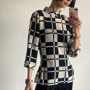 Puede incluir: Una blusa a cuadros en blanco y negro con cuello alto y mangas tres cuartos. La parte superior tiene una cintura ajustada y se combina con pantalones negros. El patrón de la blusa es un diseño geométrico audaz.