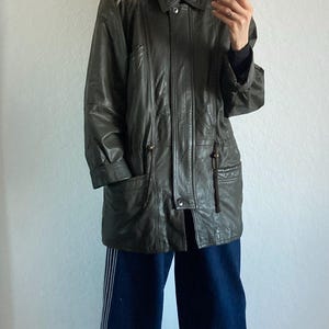 May include: Olive green leather jacket with a fur-trimmed hood. The jacket has a button-up front, pockets, and a drawstring waist. The person is wearing dark blue wide-leg pants with white stripes and gray suede shoes.
