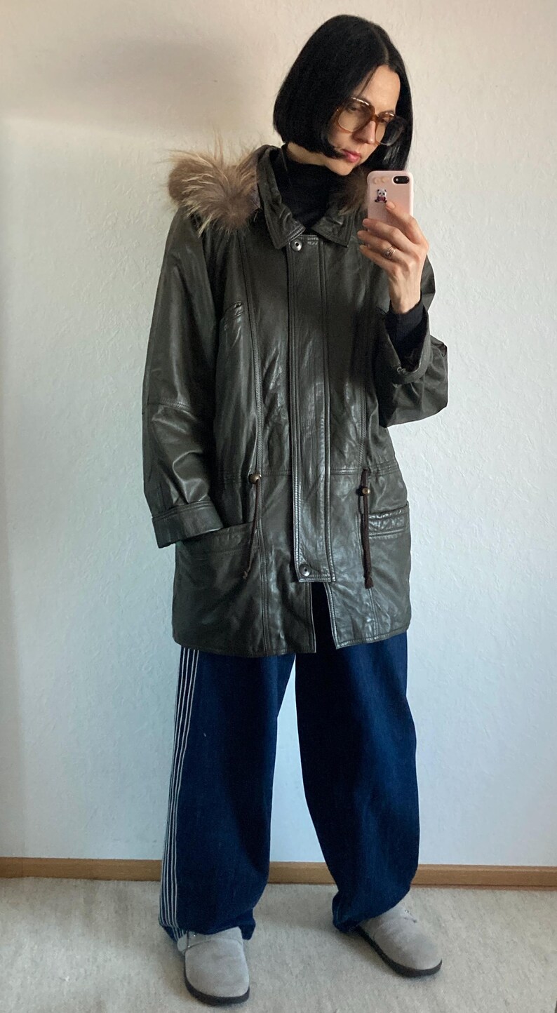 May include: Olive green leather jacket with a fur-trimmed hood. The jacket has a button-up front, pockets, and a drawstring waist. The person is wearing dark blue wide-leg pants with white stripes and gray suede shoes.
