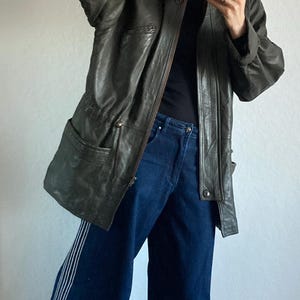 May include: Olive green leather jacket with a fur-lined hood, paired with dark blue wide-leg jeans featuring white side stripes. The outfit is completed with gray clogs.