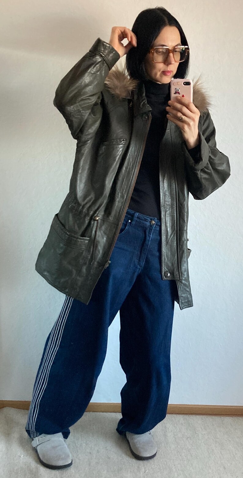 May include: Olive green leather jacket with a fur-lined hood, paired with dark blue wide-leg jeans featuring white side stripes. The outfit is completed with gray clogs.