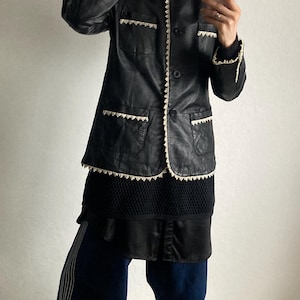 May include: Black leather jacket with cream-colored trim and pockets. The jacket is buttoned and worn over a black top and dark blue pants. The pants have a white stripe down the side and are paired with yellow sneakers.