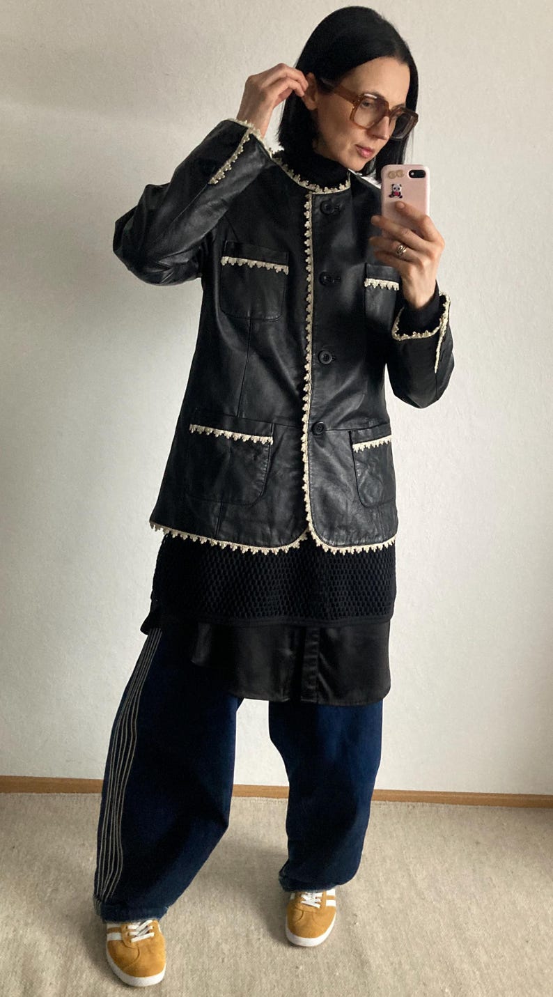 May include: Black leather jacket with cream-colored trim and pockets. The jacket is buttoned and worn over a black top and dark blue pants. The pants have a white stripe down the side and are paired with yellow sneakers.