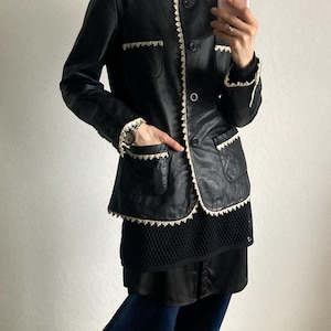 May include: Black leather jacket with cream-colored trim and button closure. The jacket has two front pockets and is worn over a black top and dark blue pants. The outfit is completed with yellow sneakers.