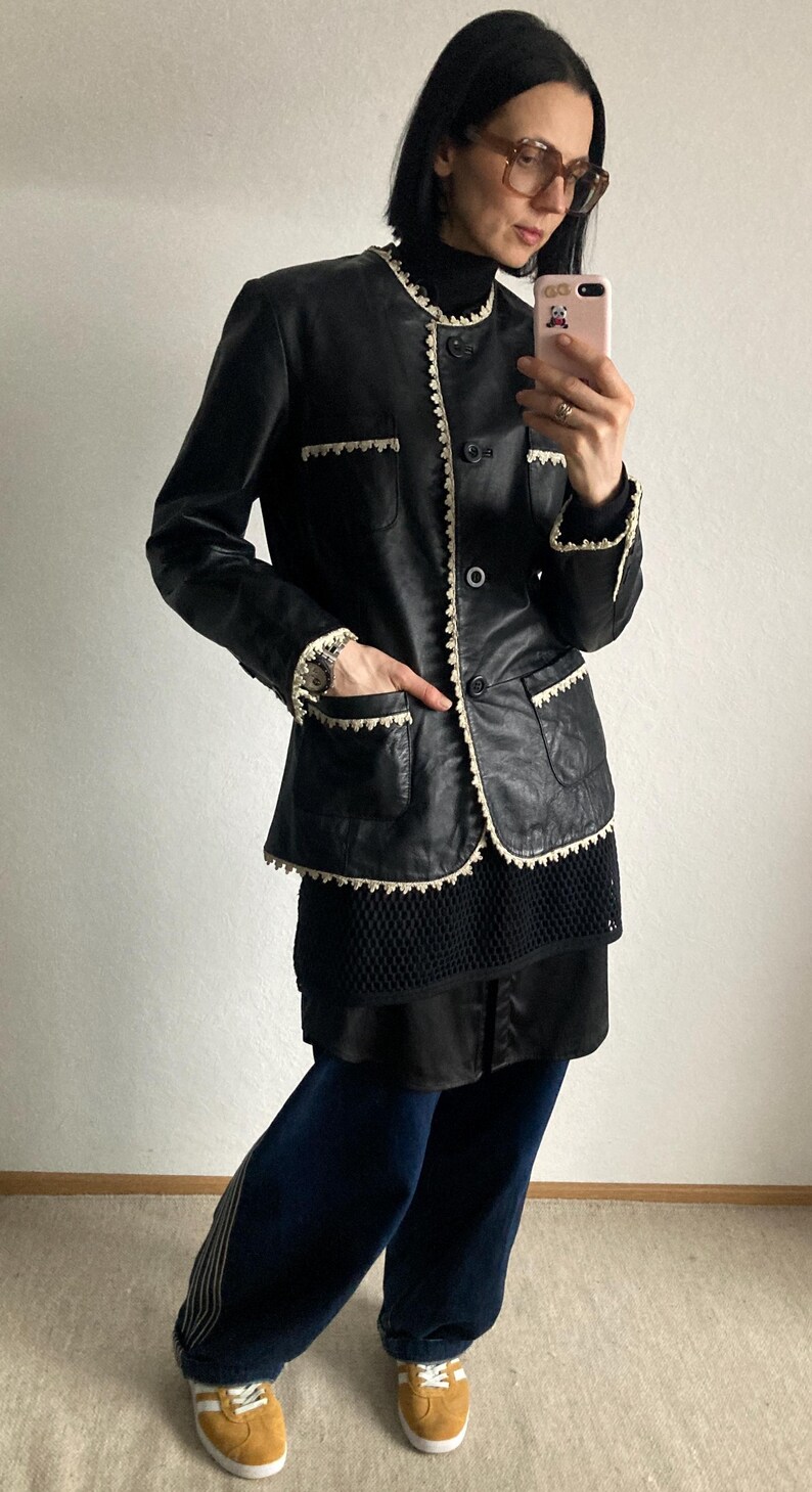 May include: Black leather jacket with cream-colored trim and button closure. The jacket has two front pockets and is worn over a black top and dark blue pants. The outfit is completed with yellow sneakers.