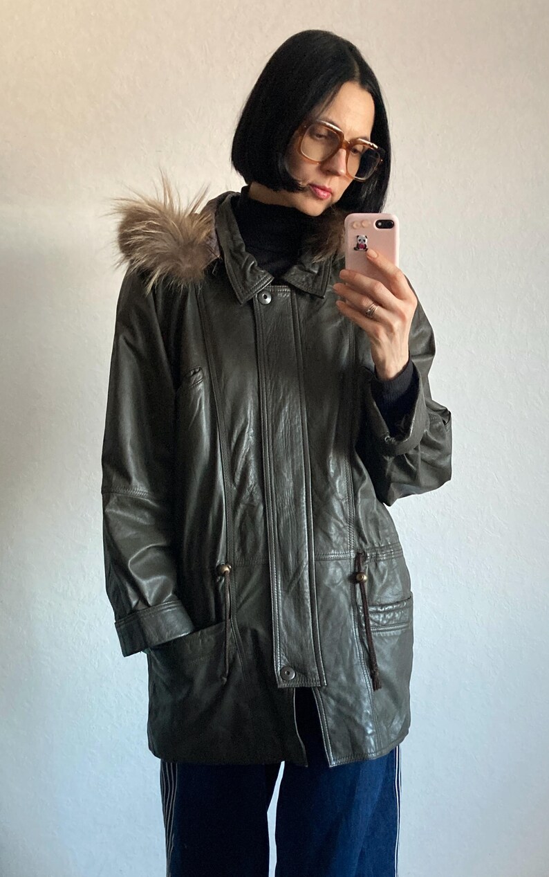 May include: Olive green leather jacket with a fur-trimmed hood. The jacket features a button-down front, pockets, and a drawstring waist. The jacket is a classic outerwear piece.