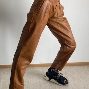 May include: Brown leather pants with a high waist and pleated front. The pants are paired with black and white sneakers with a geometric pattern. The pants have a tailored fit and a vintage aesthetic.