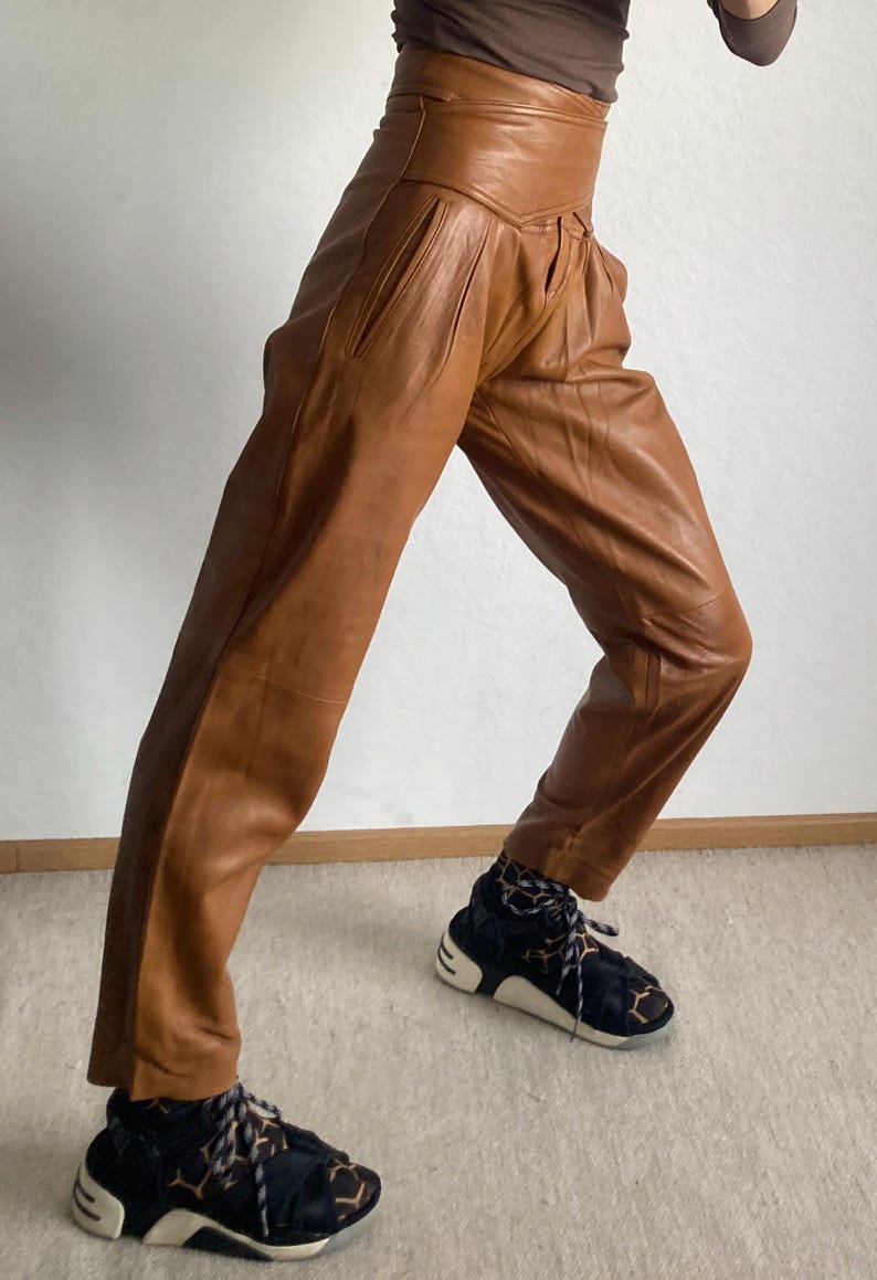May include: Brown leather pants with a high waist and pleated front. The pants are paired with black and white sneakers with a geometric pattern. The pants have a tailored fit and a vintage aesthetic.