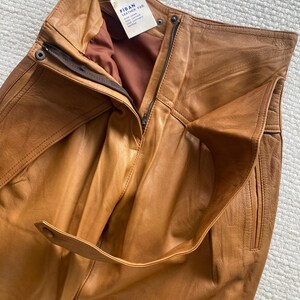 May include: Close-up of tan leather pants with a high waist and a zipper closure. The pants have a belt loop detail and a tag that reads "FIDAN LEATHER FAB". The leather appears to be soft and supple.
