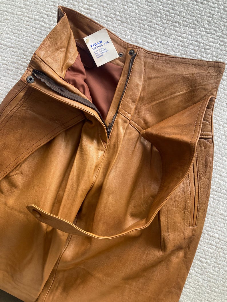 May include: Close-up of tan leather pants with a high waist and a zipper closure. The pants have a belt loop detail and a tag that reads "FIDAN LEATHER FAB". The leather appears to be soft and supple.