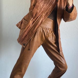 May include: A vintage brown leather jacket and matching pants set. The jacket has a classic design with pockets and a zipper. The pants are high-waisted with a pleated front. The outfit is paired with black and white patterned sneakers.