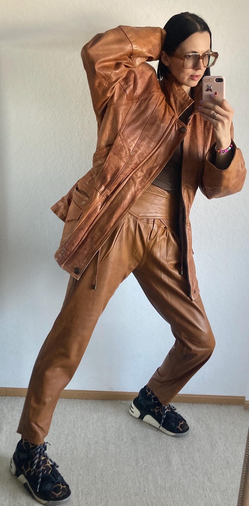 May include: A vintage brown leather jacket and matching pants set. The jacket has a classic design with pockets and a zipper. The pants are high-waisted with a pleated front. The outfit is paired with black and white patterned sneakers.
