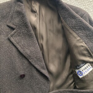 May include: A dark brown wool coat with a double-breasted design. The coat features two rows of dark brown buttons, a notched lapel, and a satin lining. The interior lining has a label that reads "First Class".