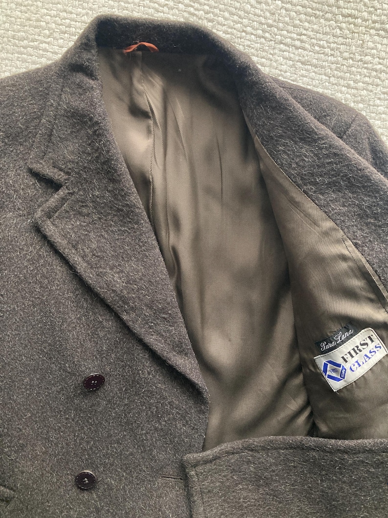 May include: A dark brown wool coat with a double-breasted design. The coat features two rows of dark brown buttons, a notched lapel, and a satin lining. The interior lining has a label that reads "First Class".