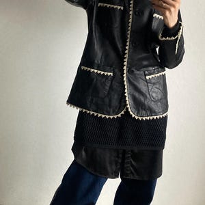 May include: Black leather jacket with white trim and a matching skirt. The jacket has a button-up front and two front pockets. The outfit is paired with dark blue pants and tan sneakers.
