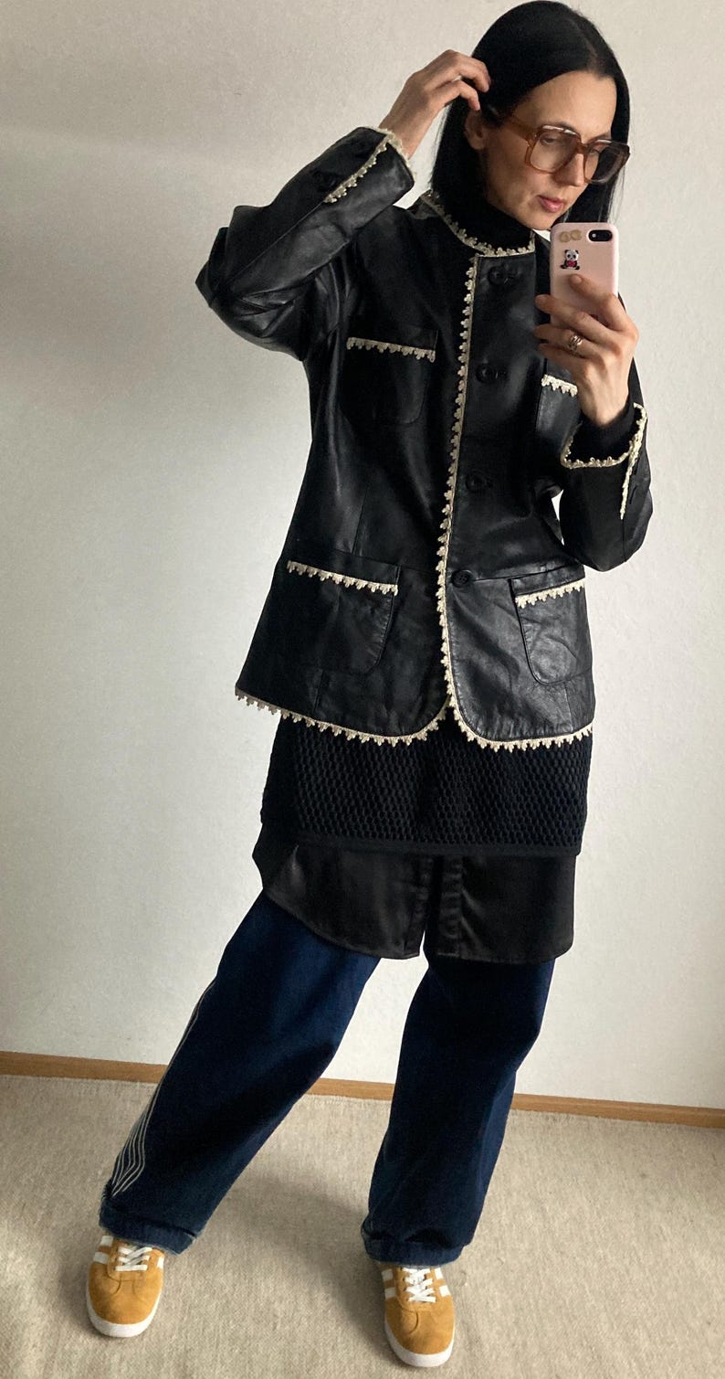 May include: Black leather jacket with white trim and a matching skirt. The jacket has a button-up front and two front pockets. The outfit is paired with dark blue pants and tan sneakers.