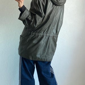 May include: A dark olive green leather jacket with a fur-lined hood. The jacket has a drawstring waist and two front pockets. The person is wearing blue jeans with white stripes down the sides.