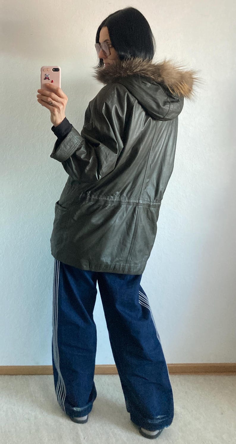 May include: A dark olive green leather jacket with a fur-lined hood. The jacket has a drawstring waist and two front pockets. The person is wearing blue jeans with white stripes down the sides.