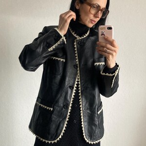 May include: Black leather jacket with cream-colored lace trim around the collar, pockets, and front closure. The jacket has a button-up front and a classic, tailored design. The jacket is worn over a black top.