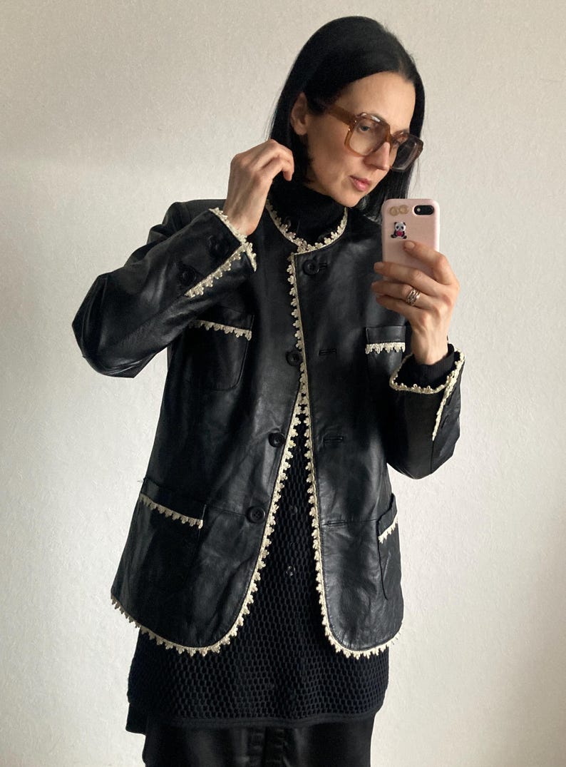 May include: Black leather jacket with cream-colored lace trim around the collar, pockets, and front closure. The jacket has a button-up front and a classic, tailored design. The jacket is worn over a black top.