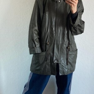 May include: A dark green leather jacket with a fur-lined hood. The jacket has a button closure, pockets, and a drawstring waist. The person is wearing blue jeans with white stripes and gray shoes. The jacket appears to be vintage.