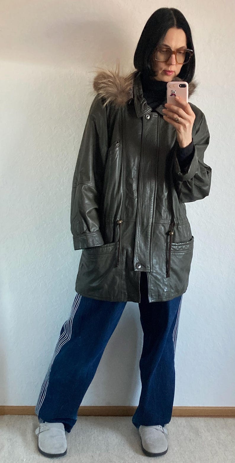 May include: A dark green leather jacket with a fur-lined hood. The jacket has a button closure, pockets, and a drawstring waist. The person is wearing blue jeans with white stripes and gray shoes. The jacket appears to be vintage.