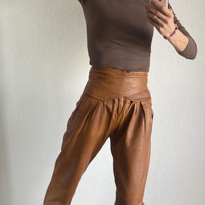 May include: A brown long-sleeve top paired with high-waisted, tan leather pants. The pants have a unique wrap-around design at the waist. The outfit is completed with black and tan patterned sneakers. The overall style is casual and chic.