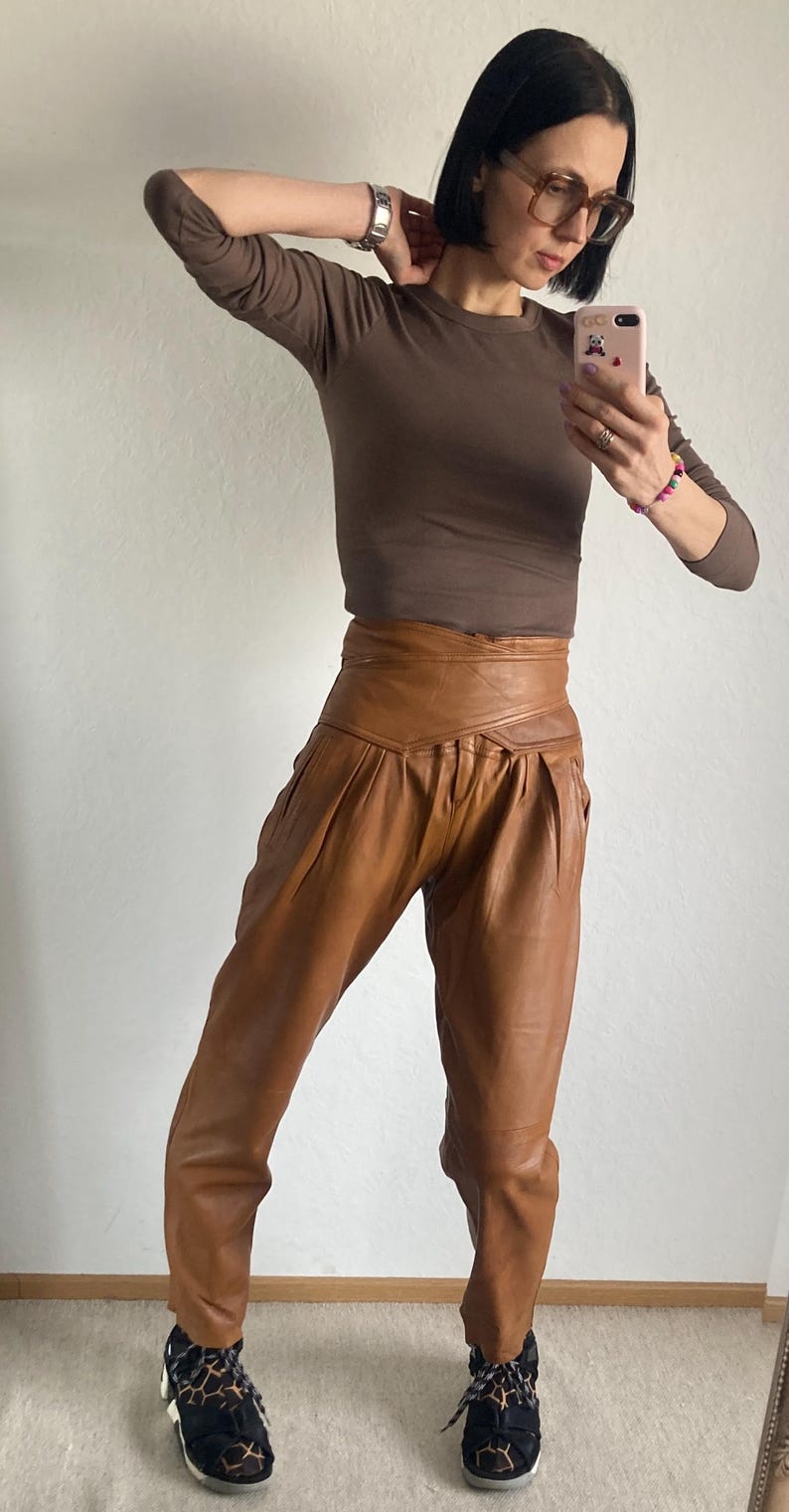 May include: A brown long-sleeve top paired with high-waisted, tan leather pants. The pants have a unique wrap-around design at the waist. The outfit is completed with black and tan patterned sneakers. The overall style is casual and chic.