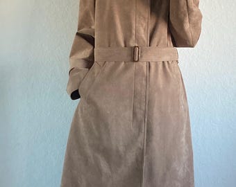 Könnte beinhalten: Ein beigefarbener Wildleder-Trenchcoat mit passendem Gürtel. Der Mantel hat einen klassischen Kragen und Taschen. Die Person trägt eine schwarze Hose mit weißen Streifen und weiße und braune Hausschuhe. Der Hintergrund ist eine schlichte weiße Wand.